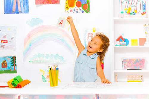 Smart little preschool girl with curly hair showing flashcard with letter A in kindergarten class with exiting emotion lifting card up